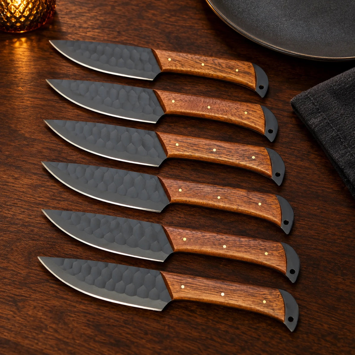 Six black steak knives with brass-pinned rosewood handles beside a plate and napkin