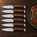 Set of 6 hammered Damascus steak knives with rosewood handles beside a grilled steak